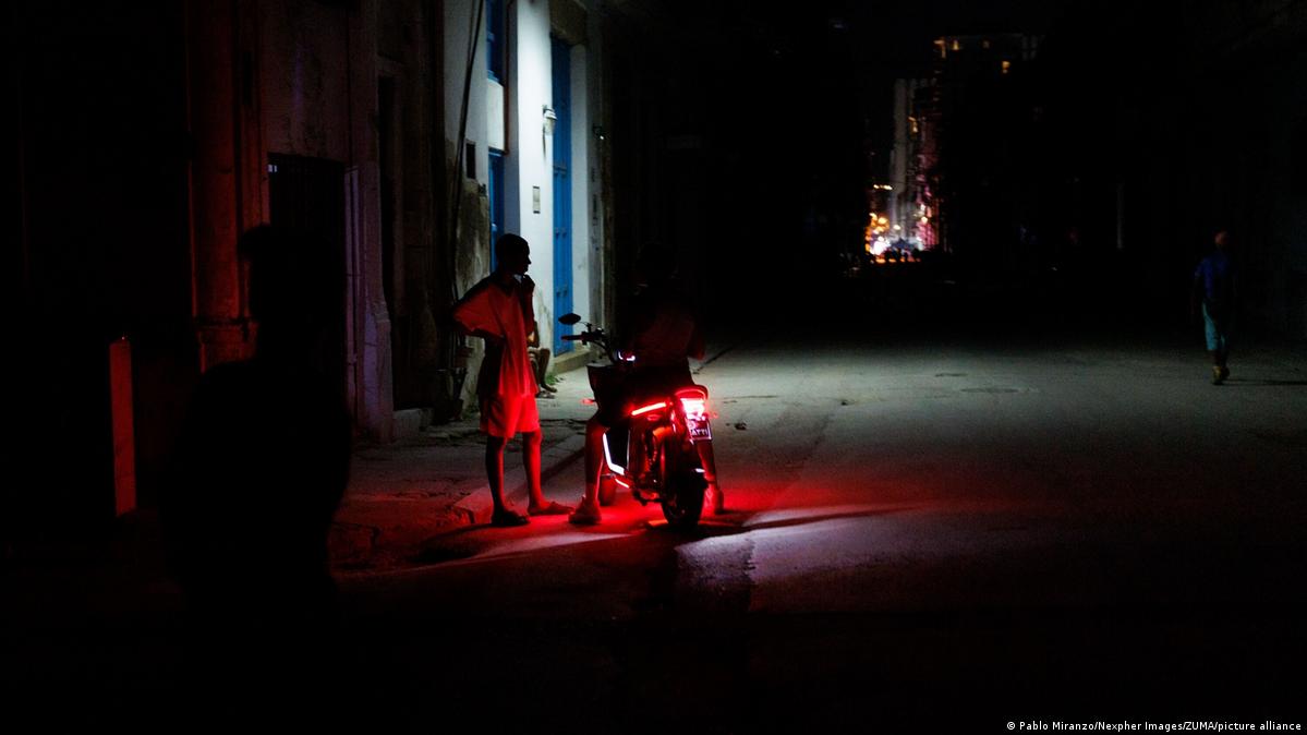 Locals stand near a motorbike with its taillight on to provide minimal illumination on an unlit street during a blackout in Central Havana, Cuba, on June 16, 2026.