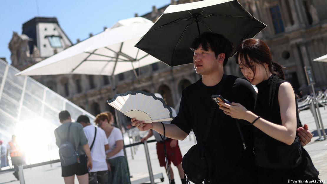Paris'teki Louvre Müzesi önünde bir kadın ve erkek, güneşten korunmak için şemsiye kullanıyor. Ön plandaki kişi elindeki vantilatörle serinlemeye çalışırken çevrede başka ziyaretçiler de görülüyor
