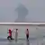 Irán, Bandar Abbas, 2026 | Niños juegan en el mar, mientras se observa humo al fondo, en el estrecho de Ormuz.