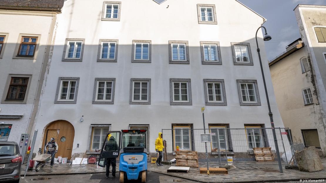 Braunau am Inn'deki binanın ön cephesindeki inşaat çalışmaları görülüyor. Giriş kapısının önünde işçiler, yapı malzemeleri, küçük bir iş makinesi ve güvenlik bariyerleri bulunuyor. Cephede çok sayıda pencere yer alıyor Braunau am Inn'deki binanın ön cephesindeki inşaat çalışmaları görülüyor. Giriş kapısının önünde işçiler, yapı malzemeleri, küçük bir iş makinesi ve güvenlik bariyerleri bulunuyor. Cephede çok sayıda pencere yer alıyor
