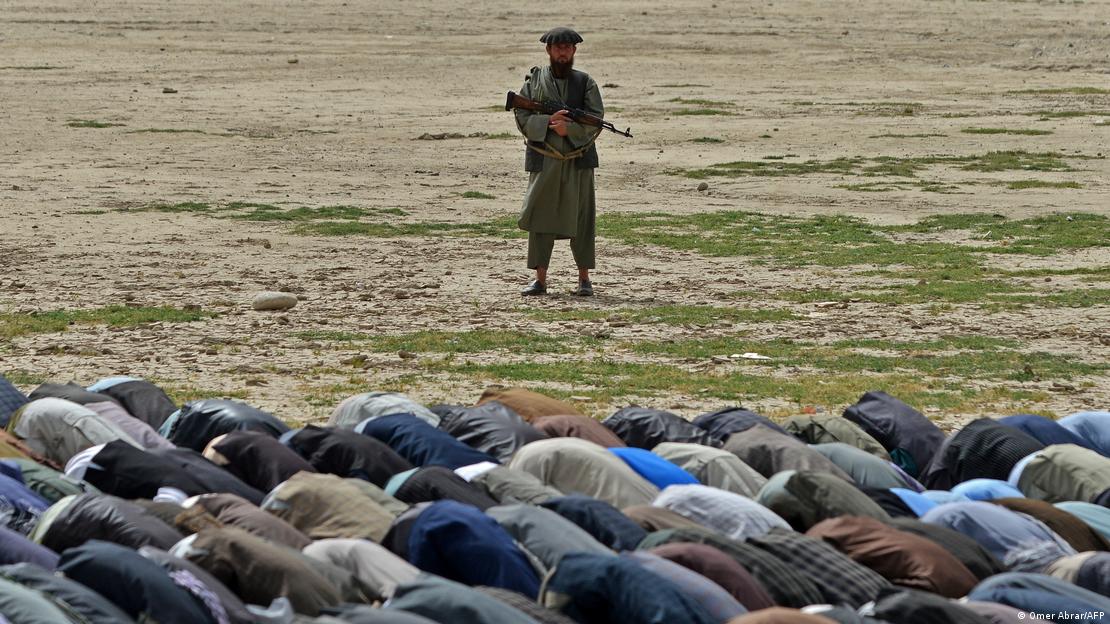 Açık arazide topluca namaz kılan, secdede insan kalabalığı ve eli silahlı bir güvenlik görevlisi