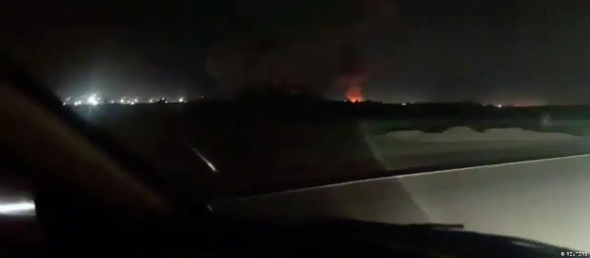 Una toma nocturna desde un vehículo mostrando lo que parecen ser explosiones y fuego a lo lejos