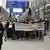 Links vorne im Bild Polizisten, im Hintergrund eine Demonstration mit vielen Menschen in einer Flughafenhalle. Sie tragen ein Banner mit der Aufschrift: "Stop deportations. Refugees welcome"