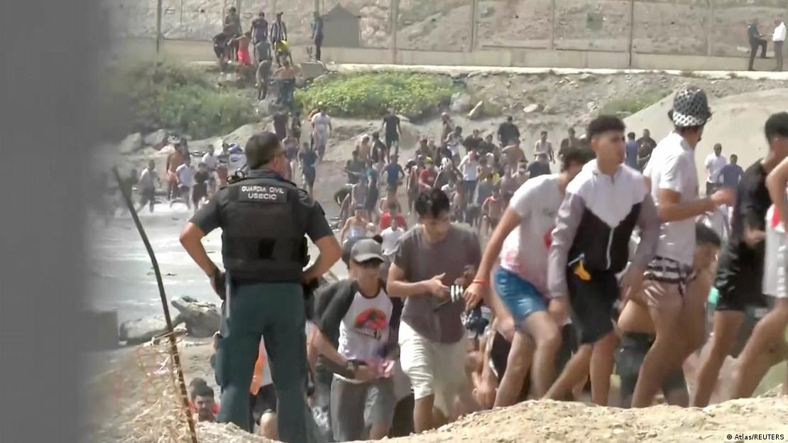 Kalabalık bir grubun sınır hattına yakın bir bölgede hareket ettiği, ön planda ise bir güvenlik görevlisinin kalabalığı izlediği görülüyor