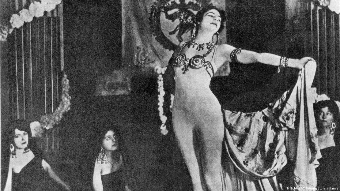 1905 yılına ait fotoğrafında Mata Hari, sahnede müzisyenlerin önünde kollarını açarak dans ediyor; üzerinde vücudunu kısmen örten dökümlü bir kostüm bulunuyor. 1905 yılına ait fotoğrafında Mata Hari, sahnede müzisyenlerin önünde kollarını açarak dans ediyor; üzerinde vücudunu kısmen örten dökümlü bir kostüm bulunuyor.