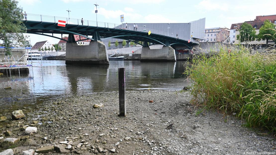 Regensburg'da Tuna Nehri'nin çekilen kıyısında taşlı nehir yatağı açığa çıkmış durumda. Arka planda nehir üzerindeki bir köprü ve daralan su yüzeyi bulunuyor.