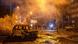 A imagem mostra destroços de um automóvel e pessoas observando a destruição em volta de uma área escura, iluminada ao fundo, com muita fumaça, devido a um ataque russo na região de Kiev, capital da Ucrânia.