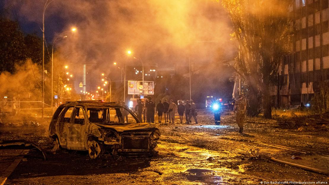 A imagem mostra destroços de um automóvel e pessoas observando a destruição em volta de uma área escura, iluminada ao fundo, com muita fumaça, devido a um ataque russo na região de Kiev, capital da Ucrânia.