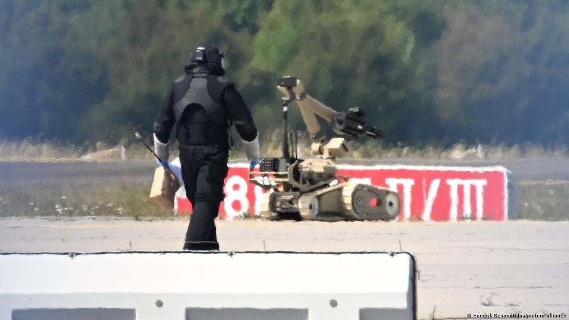 Bir havaalanı pistinde bomba imha robotu ve ona doğru yürüyen, özel kıyafetli bomba imha uzmanı Bir havaalanı pistinde bomba imha robotu ve ona doğru yürüyen, özel kıyafetli bomba imha uzmanı