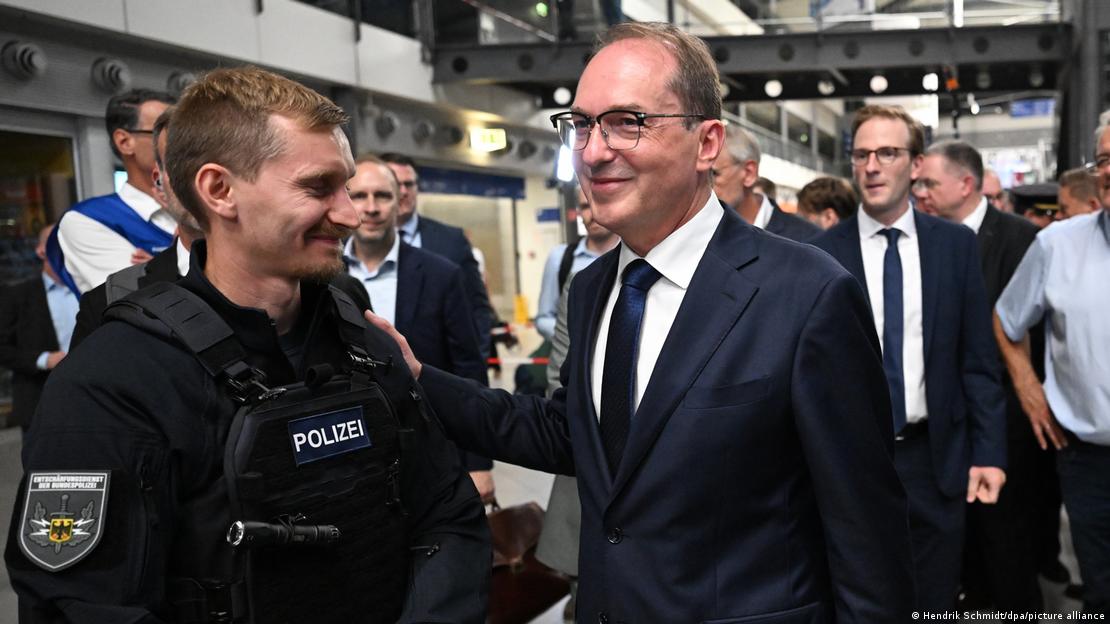 Almanya İçişleri Bakanı Alexander Dobrindt, bir polisin omuzuna gülümseyerek dokunuyor, etrafta yetkililer ve görevlilerden oluşan bir kalabalık var Almanya İçişleri Bakanı Alexander Dobrindt, bir polisin omuzuna gülümseyerek dokunuyor, etrafta yetkililer ve görevlilerden oluşan bir kalabalık var