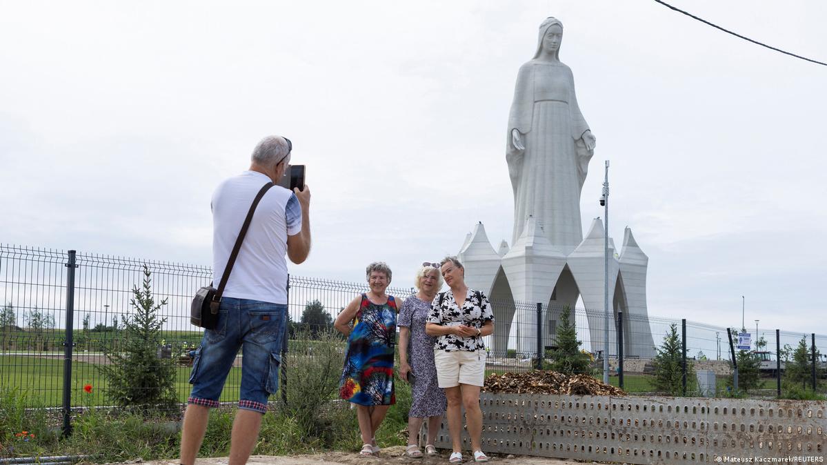 A man takes a photo of three smiling women in front of a huge white statue of the Virgin Mary, Konotopie, Poland, July 29, 2026