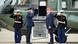 Donald Trump, in a dark blue suit and red, peaked MAGA cap, stands side on in front of the steps up into a fighter aircraft with "Welcome Aboard Marine One" written on them. Three men in formal uniforms are saluting him.