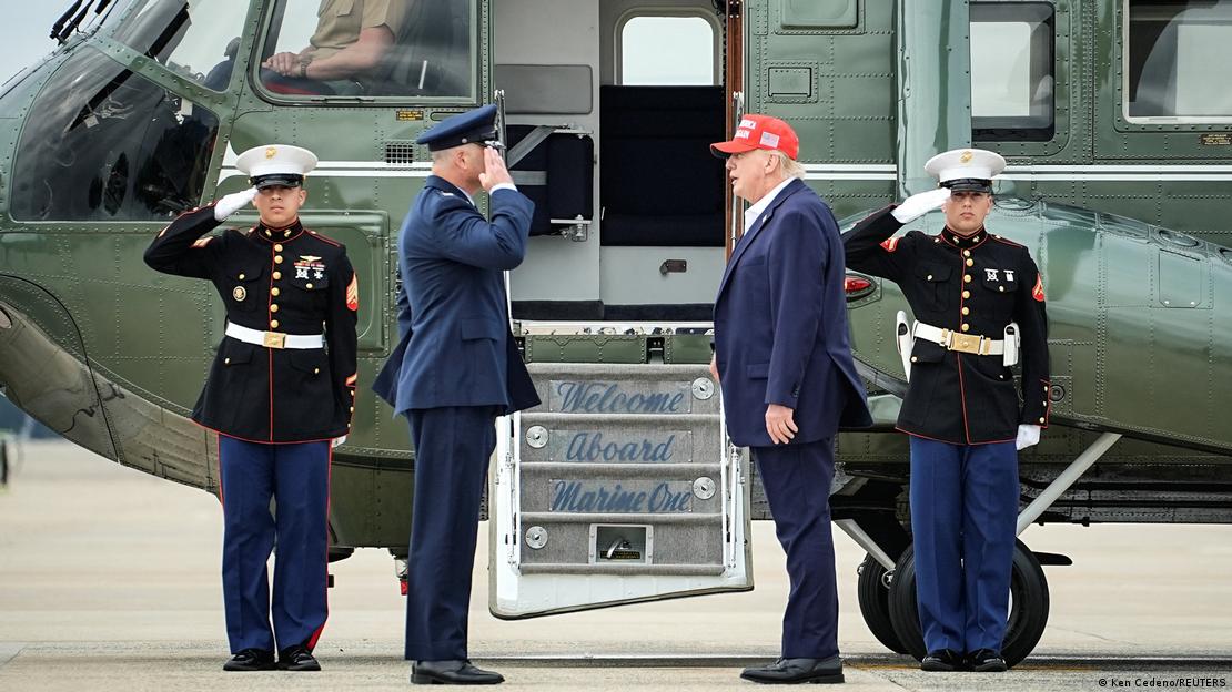 Donald Trump, in a dark blue suit and red, peaked MAGA cap, stands side on in front of the steps up into a fighter aircraft with "Welcome Aboard Marine One" written on them. Three men in formal uniforms are saluting him. Donald Trump, in a dark blue suit and red, peaked MAGA cap, stands side on in front of the steps up into a fighter aircraft with "Welcome Aboard Marine One" written on them. Three men in formal uniforms are saluting him.
