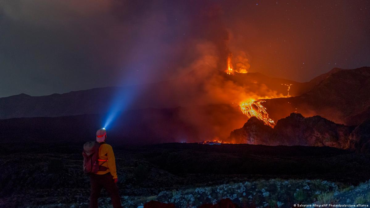 A person with a head lamp stands in the darkness in front of glowing orange rivers of lava expelled from a fountain, while a plume of smoke can be seen behind (13.08.2026)