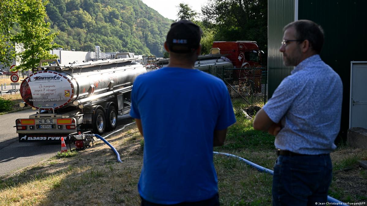 Two men, dressed in shades of blue, stand outside near two silver tanker trucks filled with water