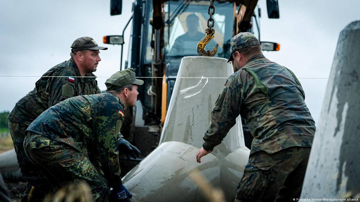 Three soldiers in camouflage uniforms and peaked caps stand around a big Y-shaped concrete block. In the background, a man sits in the cabin of a backhoe loader