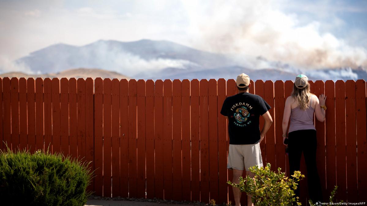 Two people look over a fence at a fire burning in the hills