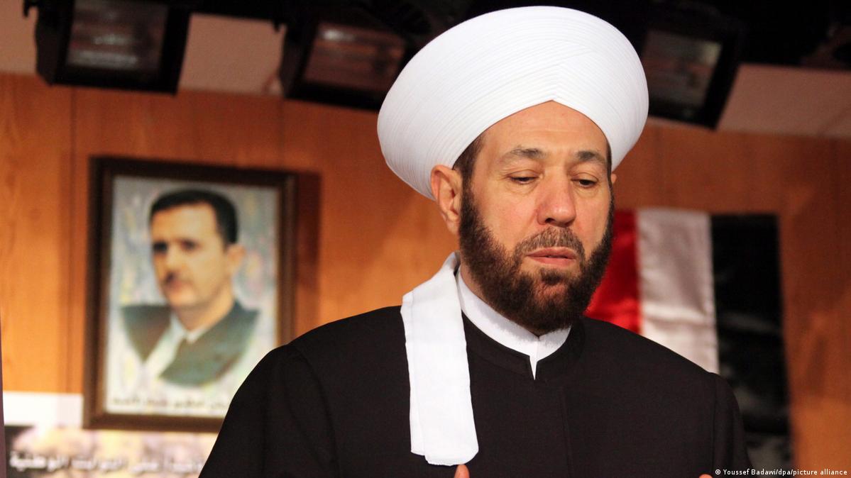 Ahmad Hassoun in a white head dressing with a blurred picture of Bashar Assad behind him