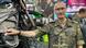 A man in camouflage shirt with a hand on an army motorcycle.