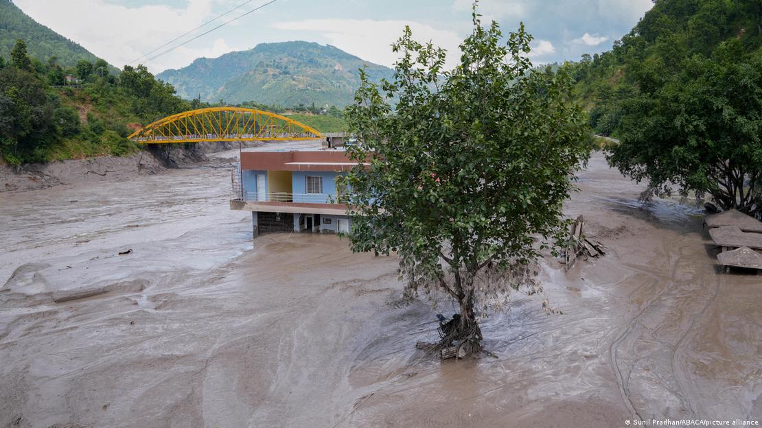 Nepal'in Nuwakot bölgesinde ani selin ardından bir ev, çevresini kaplayan sel suyu ve çamurun ortasında kalmış durumda; arka planda köprü ve dağlık alan yer alıyor