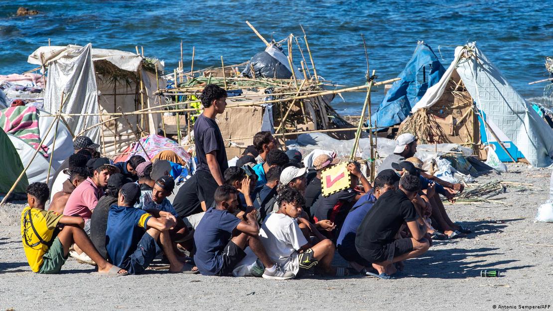 Imigrantes esperam por distribuição de alimentos em praia de Ceuta; ao fundo, barracos improvisados