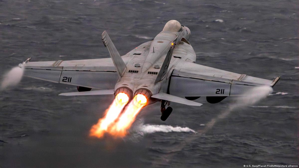 A grey fighter aircraft, seen from behind, flying low over the sea with two jets of flame shooting out from the back.