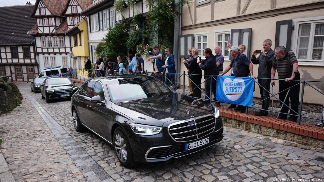 Blick in eine Straße mit vielen Fachwerkhäusern: Im Vordergrund fährt ein schwarzes Fahrzeug mit Berliner Kennzeichen, dem weitere folgen. Am Straßenrand stehen mehrere Demonstranten, von denen einige ein Transparent halten mit der Aufschrift: "Unser Land zuerst" Blick in eine Straße mit vielen Fachwerkhäusern: Im Vordergrund fährt ein schwarzes Fahrzeug mit Berliner Kennzeichen, dem weitere folgen. Am Straßenrand stehen mehrere Demonstranten, von denen einige ein Transparent halten mit der Aufschrift: "Unser Land zuerst"