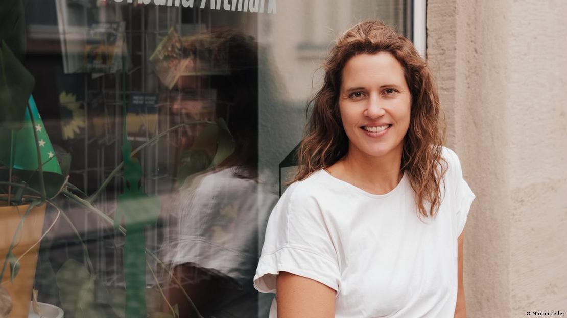 Eine Frau im weißen T-Shirt mit langen lockigen Haare, Miriam Zeller, Kandidatin der Grünen in Stendal, Miriam Zeller, steht draußen vor der Fensterscheibe des Wahlkreisbüros Eine Frau im weißen T-Shirt mit langen lockigen Haare, Miriam Zeller, Kandidatin der Grünen in Stendal, Miriam Zeller, steht draußen vor der Fensterscheibe des Wahlkreisbüros