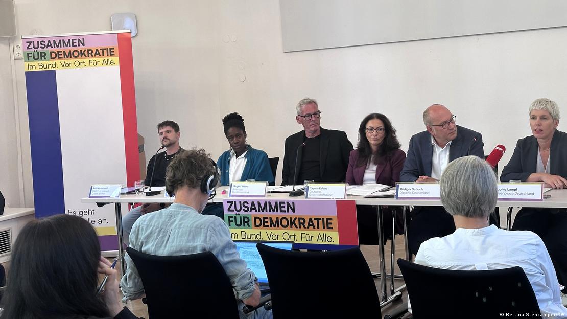 Drei Männer und drei Frauen sitzen hinter einem Tisch. Neben ihnen auf einer Stellwand und auf einem Transparent an der Tischkante steht "Zusammen für Demokratie. Im Bund. Vor Ort. Für Alle." Auf Stühlen vor ihrem Tisch sitzen mehrere Menschen im Publikum, die man nur von hinten sieht. Drei Männer und drei Frauen sitzen hinter einem Tisch. Neben ihnen auf einer Stellwand und auf einem Transparent an der Tischkante steht "Zusammen für Demokratie. Im Bund. Vor Ort. Für Alle." Auf Stühlen vor ihrem Tisch sitzen mehrere Menschen im Publikum, die man nur von hinten sieht.