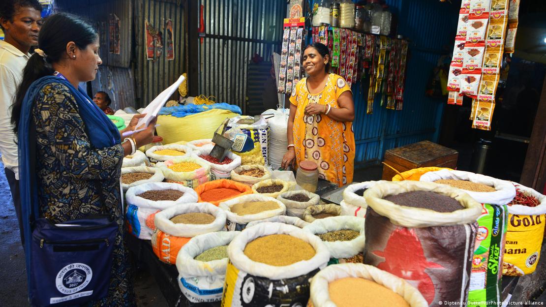 Mulher com prancha na mão entrevista vendedora de temperos em mercado na Índia