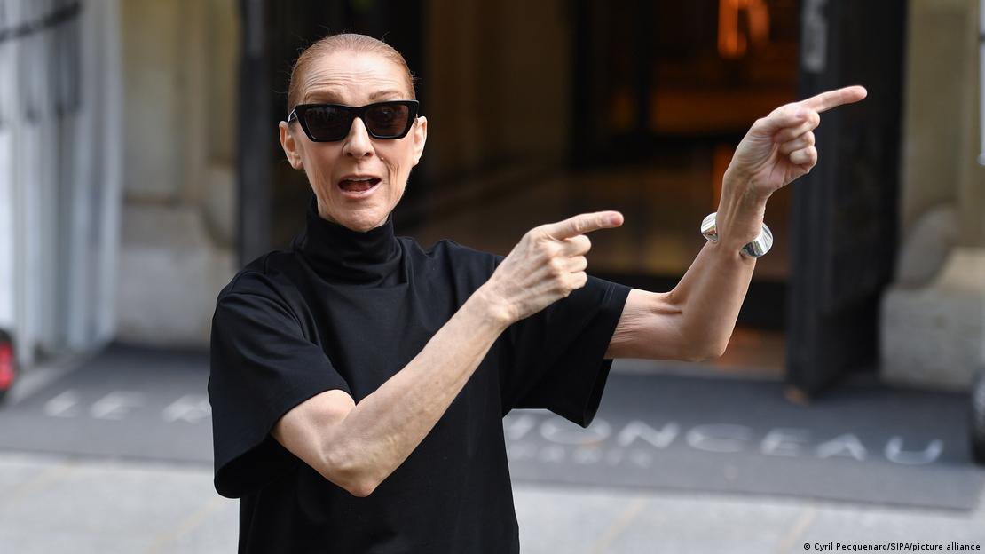 A cantura Céline Dion vestida de preto e de óculos escrutos gesticula em frente ao Hotel Royal Monceau, em Paris.