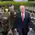 Donald Trump walking in a blue suit and red striped tie alongside two army officers, one with raised sword, with other soldiers lined up in background