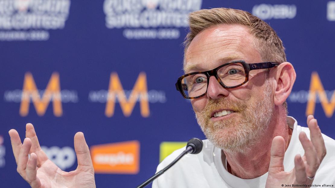 Eurovision Direktörü Martin Green, bir basın toplantısında mikrofon önünde konuşurken iki eliyle jest yapıyor. Eurovision Direktörü Martin Green, bir basın toplantısında mikrofon önünde konuşurken iki eliyle jest yapıyor.