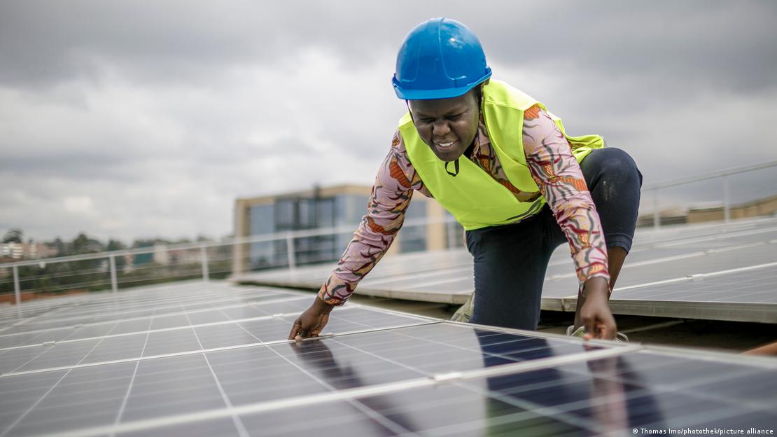 A foto mostra uma estudante da Strathmore University, no Quênia, em cima de um telhado com paineis solares. Ela é uma mulher preta, usa capacete de segurança de cor azul e um colete de segurança amarelo neon. A foto mostra uma estudante da Strathmore University, no Quênia, em cima de um telhado com paineis solares. Ela é uma mulher preta, usa capacete de segurança de cor azul e um colete de segurança amarelo neon.
