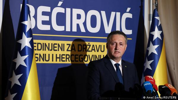 A man (Denis Becirovic) standing in front of a blue-and yellow backdrop bearing his name and between two Bosnian flags, addresses the press in Sarajevo, Bosnia and Herzegovina, October 4, 2026