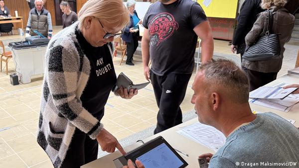 A woman points at an electronic device as a man looks on. In the background are people and what looks like a large ballot box. Knezica, Bosnia and Herzegovina, October 4, 2026