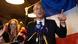 Raphael Glucksmann, a 46-year-old white man in a dark suit, pointing a finger as he speaks into a microphone in front of a blue-white-and-red French flag. In the background a young woman applauds at the front of a crowd of people. Raphael Glucksmann, a 46-year-old white man in a dark suit, pointing a finger as he speaks into a microphone in front of a blue-white-and-red French flag. In the background a young woman applauds at the front of a crowd of people.