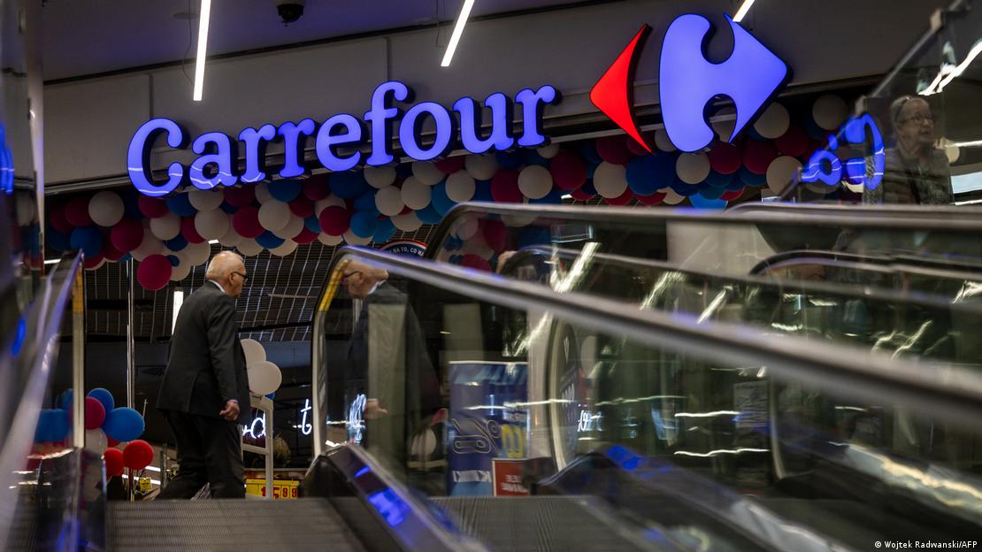 Carrefour mağazasında şirket logosu önünde yürüyen merdivenler ve müşteriler görülüyor.