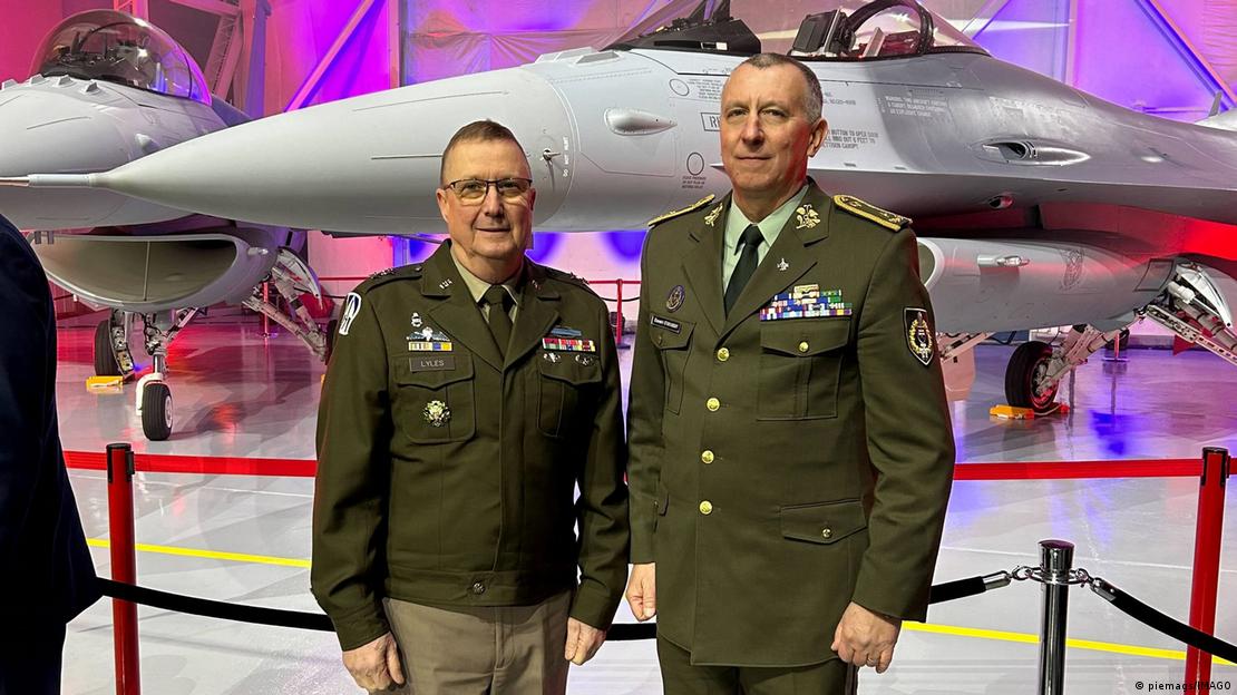 Two men (Dale Lyles and Slavomir Staviarsky) stand in front of an F-16 fighter jet in an indoor setting, Greenville, USA, February 29, 2024