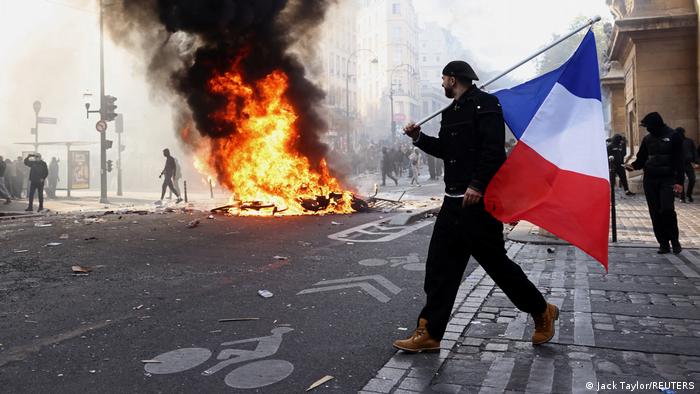 Un manifestante sostiene una bandera francesa junto a objetos en llamas en París, en medio de enfrentamientos durante una jornada de manifestaciones contra el costo de vida (17.10.2026)