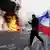 Un manifestante sostiene una bandera francesa junto a objetos en llamas en París, en medio de enfrentamientos durante una jornada de manifestaciones contra el costo de vida (17.10.2026)