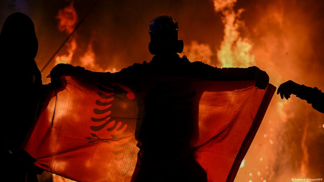 Një protestues në Tiranë ka shpalosur flamurin para një dyqani që i është vënë flaka