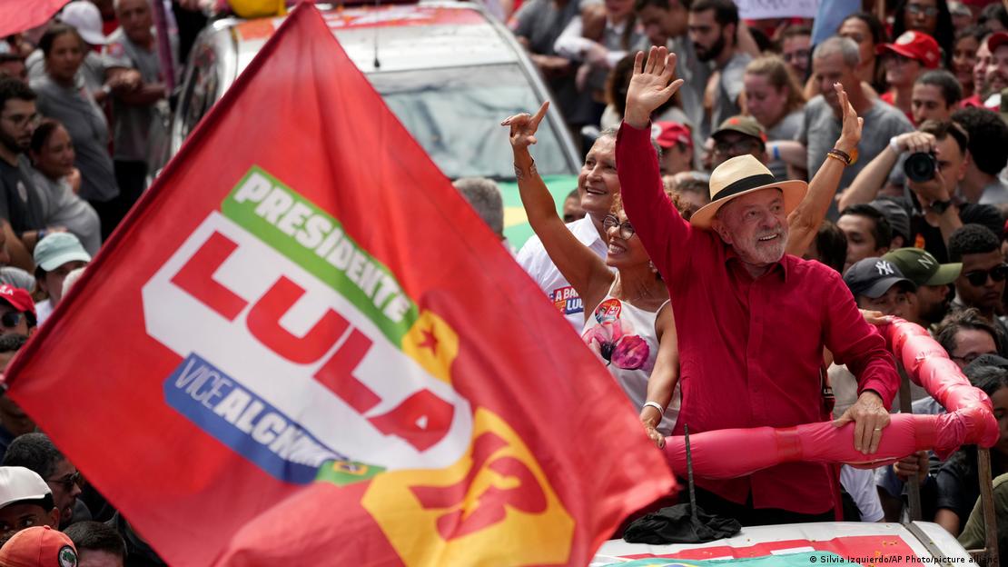 Lula da Silva, kalabalık bir seçim mitinginde aracın üzerinden el sallıyor; ön planda Lula yazılı kampanya bayrağı var