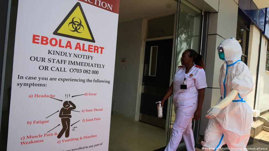 Una persona con traje de protección biológica y una enfermera frente a un cartel de alerta de ébola